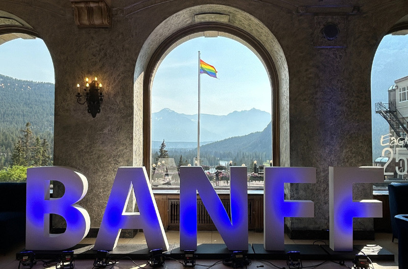 Large 3D letters lit with blue light spell “BANFF” inside an elegant room with arched stone windows. Through the central window, a rainbow Pride flag flies proudly against a clear blue sky, with pine-covered mountains and a scenic valley in the background.