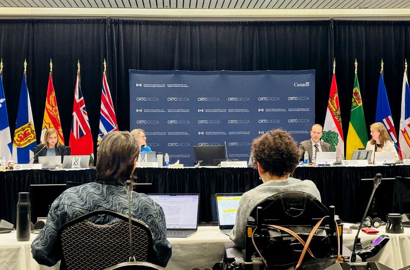 Yazmine Laroche and Winnie Luk are seen presenting at a CRTC hearing. They sit at a table facing the CRTC panel and a row of provincial flags. Yazmine uses a power wheelchair.