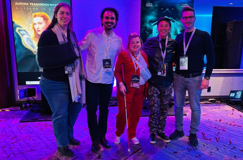 A group of five people poses in front of a vibrant backdrop featuring a screen with the title Aurora Teagarden Mysteries. From left to right: Kássia Neves (wearing an arm sling and a scarf), Amir Zargara (in a white shirt), Alicia Grace Chenier (dressed in a red jumpsuit and holding a white cane), Winnie Luk (wearing a black leather jacket and patterned pants), and Sylvain Gélinas (in a black sweater). The setting is a bright conference room with a carpeted floor.