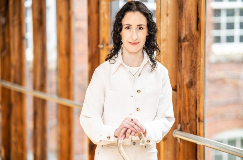 Alethea Bakogeorge stands in a sunlit wooden hallway with large windows overlooking a brick building. She wears a stylish cream-colored jacket with gold buttons, accessorized with delicate gold necklaces. Her dark curly hair frames her face, and she has a warm, confident expression. She holds a clear cane with both hands, positioned in front of her.