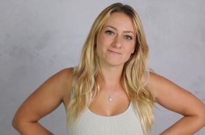 A blonde woman with long hair standing with both hands on her hips, wearing a beige tank top against a neutral background.