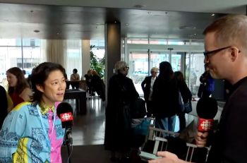 <p>Winnie Luk wearing a brightly patterned shirt is being interviewed by a CBC reporter in a busy lobby area.</p>