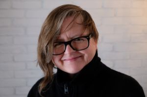Portrait d'une femme souriante aux cheveux bruns asymétriques, portant des lunettes noires et un pull noir, debout devant un fond de briques blanches.