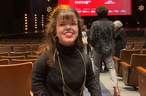 A white woman with dwarfism, with long brown hair and bangs, wears a black turtleneck, red pants, and a long gold necklace. She has red lipstick and is smiling enthusiastically at the camera.