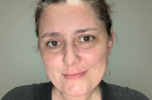 Headshot of a smiling woman with brown hair tied back, silver dangling earrings, and a black t-shirt on a gray background.